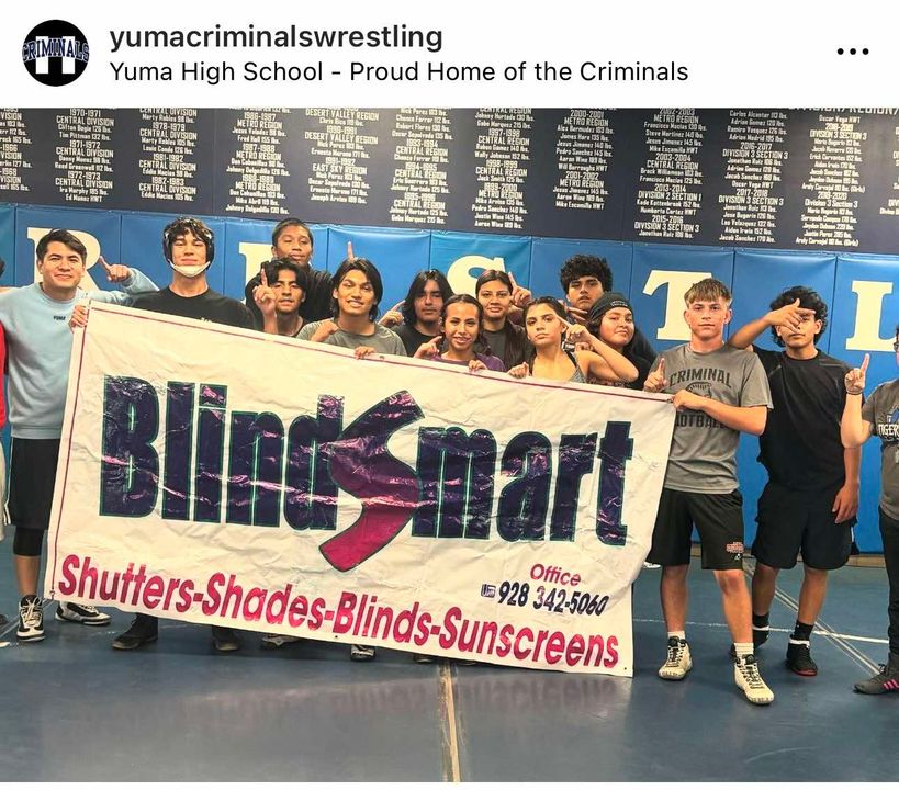 A social media screenshot from 'yumacriminalswrestling' featuring a group of student-athletes posing with a 'Blindsmart' sponsorship banner inside a gymnasium. A diverse group of teenage boys and girls stand behind a large white banner with the prominent, stylized 'Blindsmart' logo in purple and magenta. The bottom of the banner lists services: 'Shutters - Shades - Blinds - Sunscreens' with the office phone number 928 342-5060. Multiple students are pointing their index finger upwards. The background shows a large, blue wrestling mat wall with dozens of historical records and champion names printed on it. The header text reads: 'Yuma High School - Proud Home of the Criminals'.
